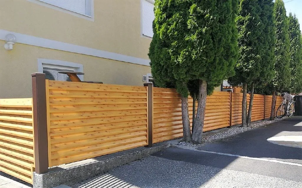 Lamellenzaun in Holzoptik entlang einer Einfahrt mit Thujen