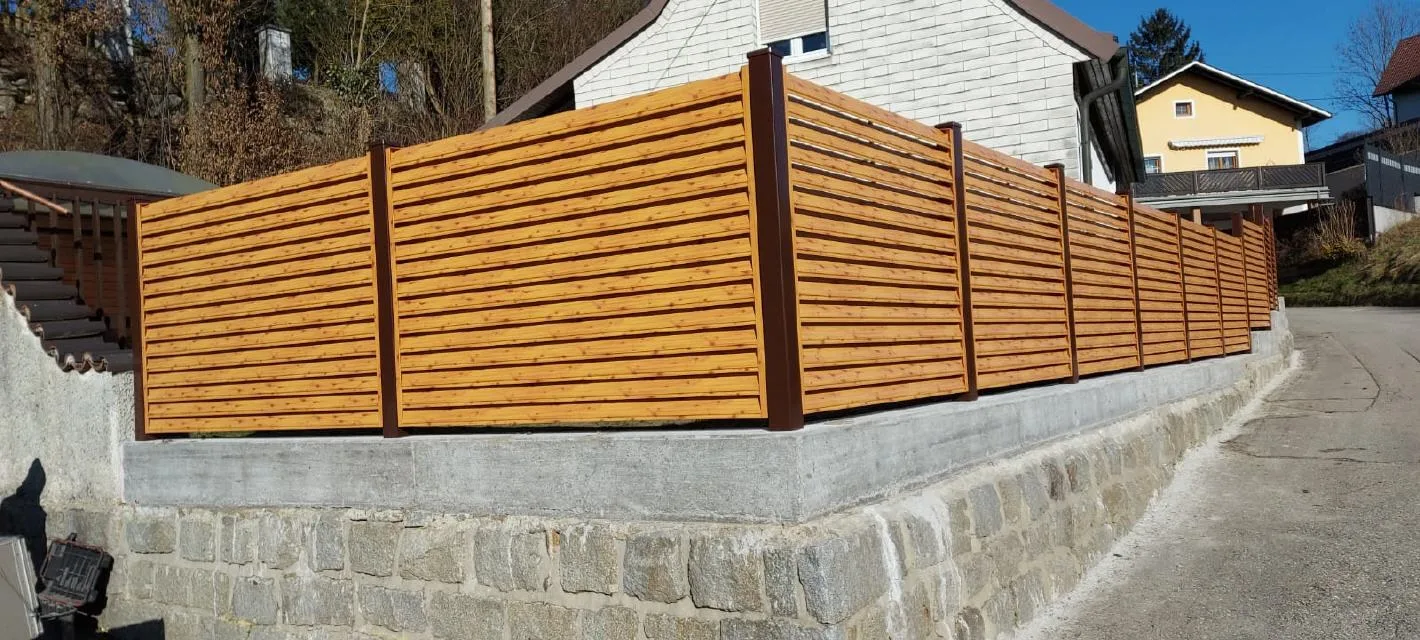 Lamellenzaun in Holzoptik auf einer Steinmauer an einer ansteigenden Straße