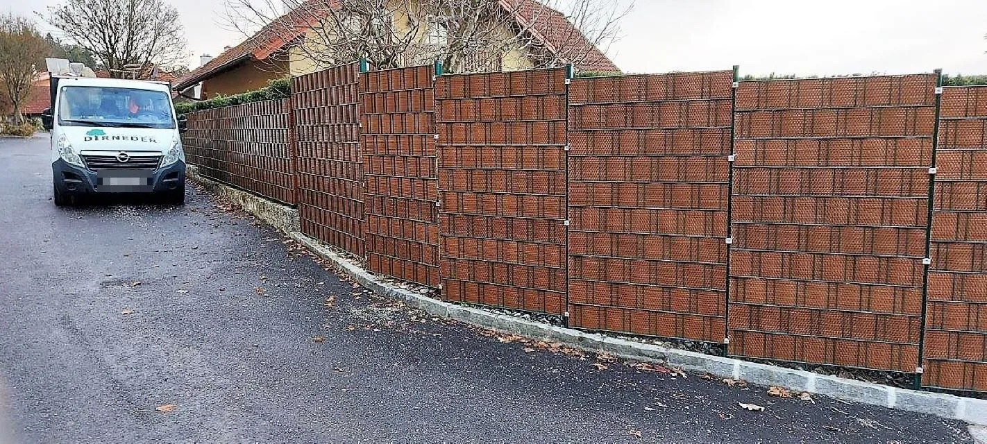 Langer brauner Sichtschutzzaun an einer Straße, links ein weißer Dirneder-Transporter